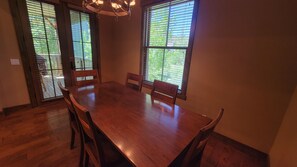 A dining area with a wooden table and chairs, large windows with blinds, and a ceiling light fixture.