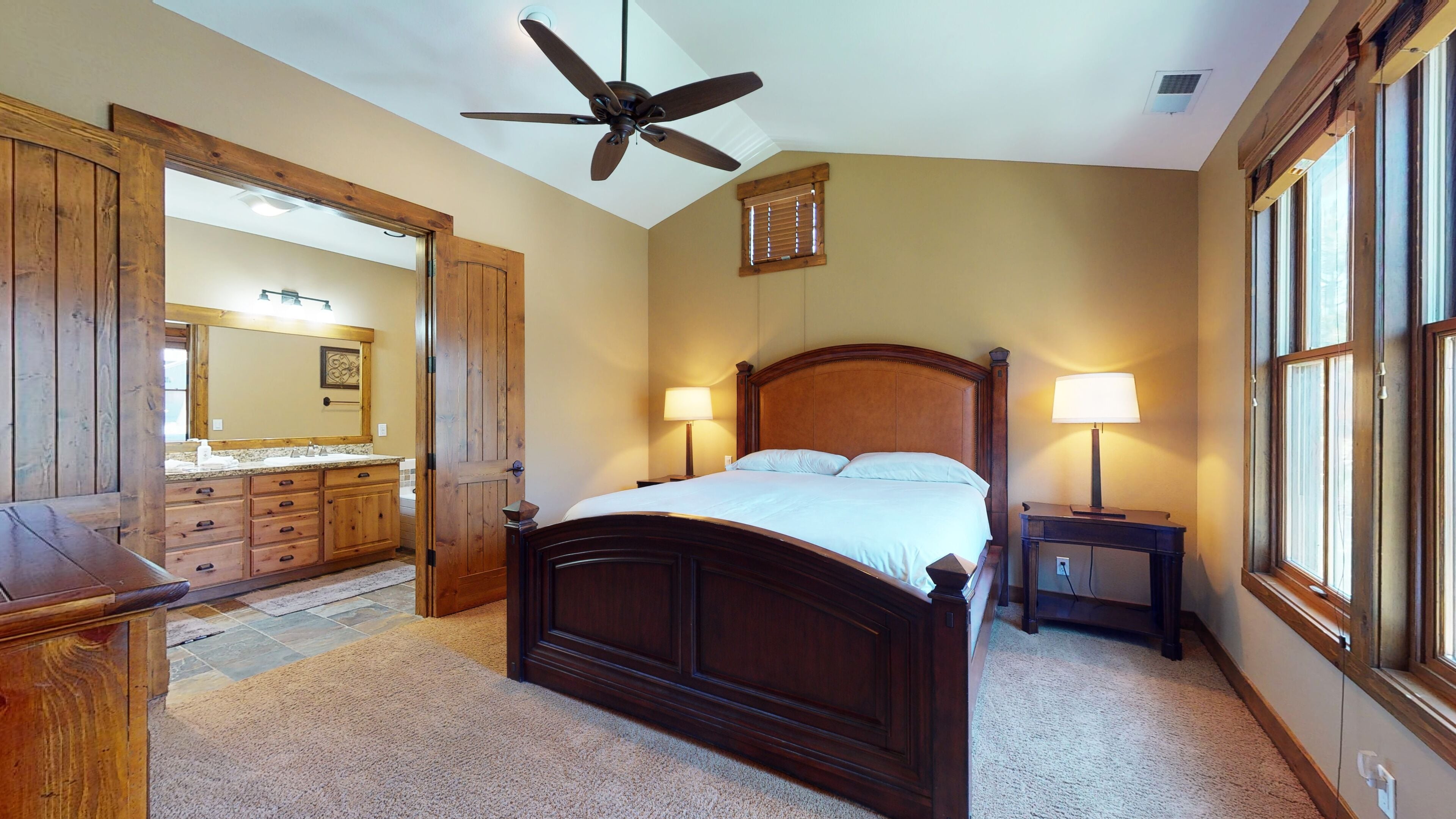 A bedroom with a wooden bed, a ceiling fan, a nightstand with a lamp, a dresser, and a large mirror.