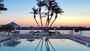 A swimming pool with lounge chairs and palm trees, surrounded by a body of water and a sunset sky.