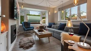 Een gezellige woonkamer met een open haard, een blauwe fauteuil, een houten salontafel en een bank met blauwe kussens. Grote ramen bieden uitzicht op het omliggende landschap.