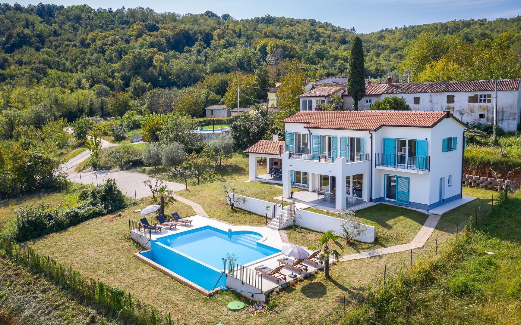 A two-story house with a swimming pool, surrounded by greenery and hills.
