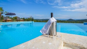 Ein klares, blaues Schwimmbecken mit einer Champagnerflasche auf einem Tisch, umgeben von Liegestühlen und einer reizvollen Hügellandschaft.