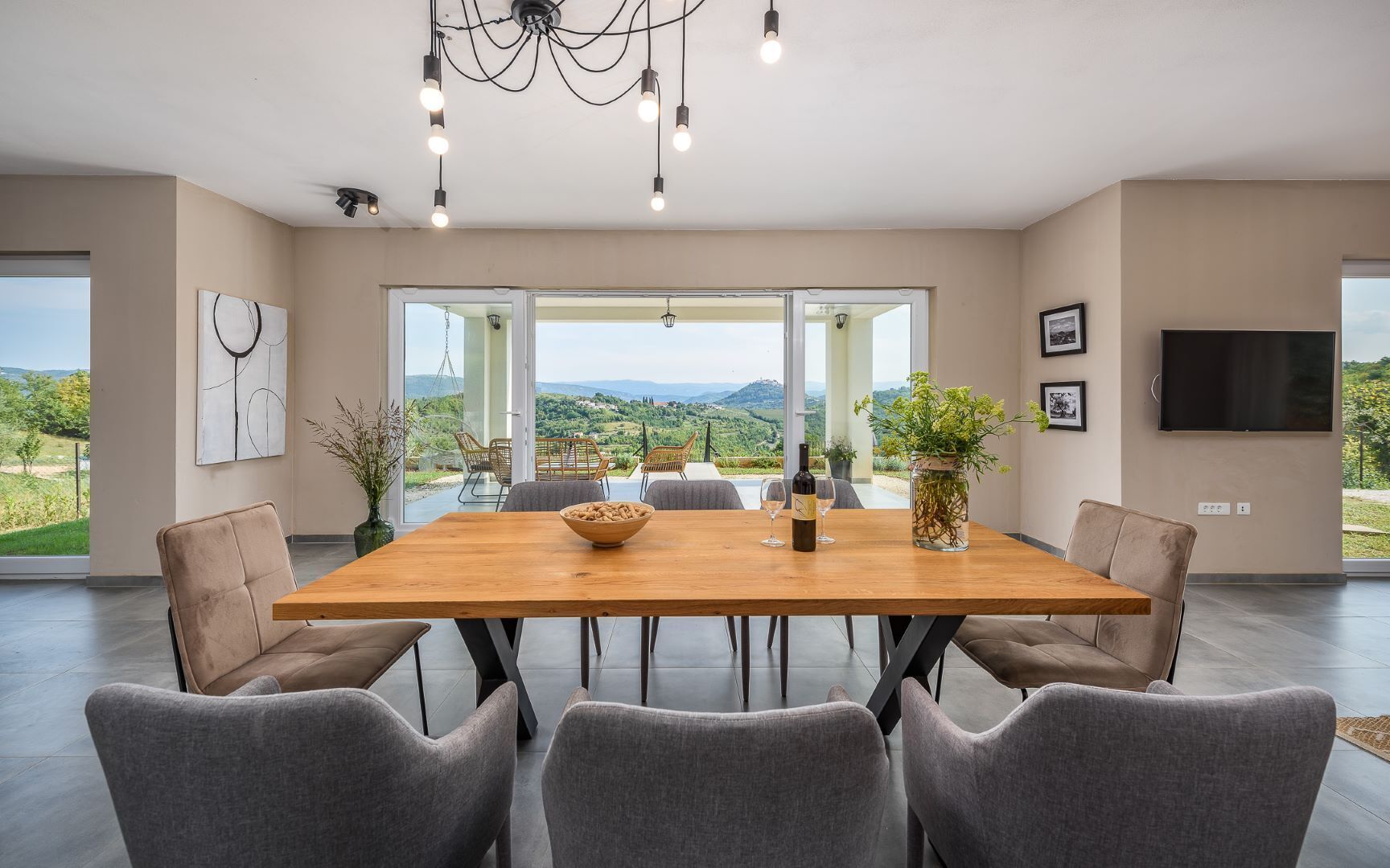 Une salle à manger spacieuse avec une grande table en bois, six chaises grises à revêtement rembourré, un lustre et une vue sur la campagne grâce à de grandes fenêtres.