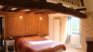 Ein Schlafzimmer mit Holzbalkendecke, einem Bett mit bunter Decke, zwei Nachttischlampen und einem Fenster mit Blick ins Freie.