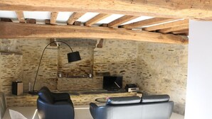 Ein gemütlicher Wohnbereich mit Steinwand, Holzbalken, einem schwarzen Ledersofa, einem grauen Sofa, einem weißen Tisch und einem Fernseher.