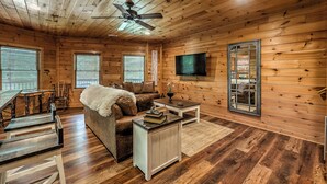 Ein gemütliches Blockhaus-Interieur mit Holzwänden, Deckenventilator, Wand-Fernseher, einer Couch, einem Couchtisch und einem Essbereich mit Stühlen und Tisch.