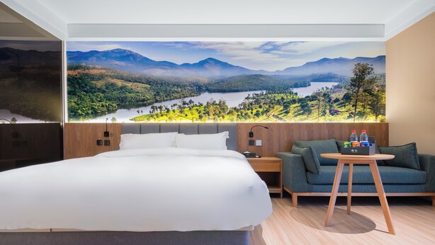 Una habitación de hotel moderna con una cama grande, un sofá verde, una pequeña mesa de madera y un mural de un paisaje montañoso.