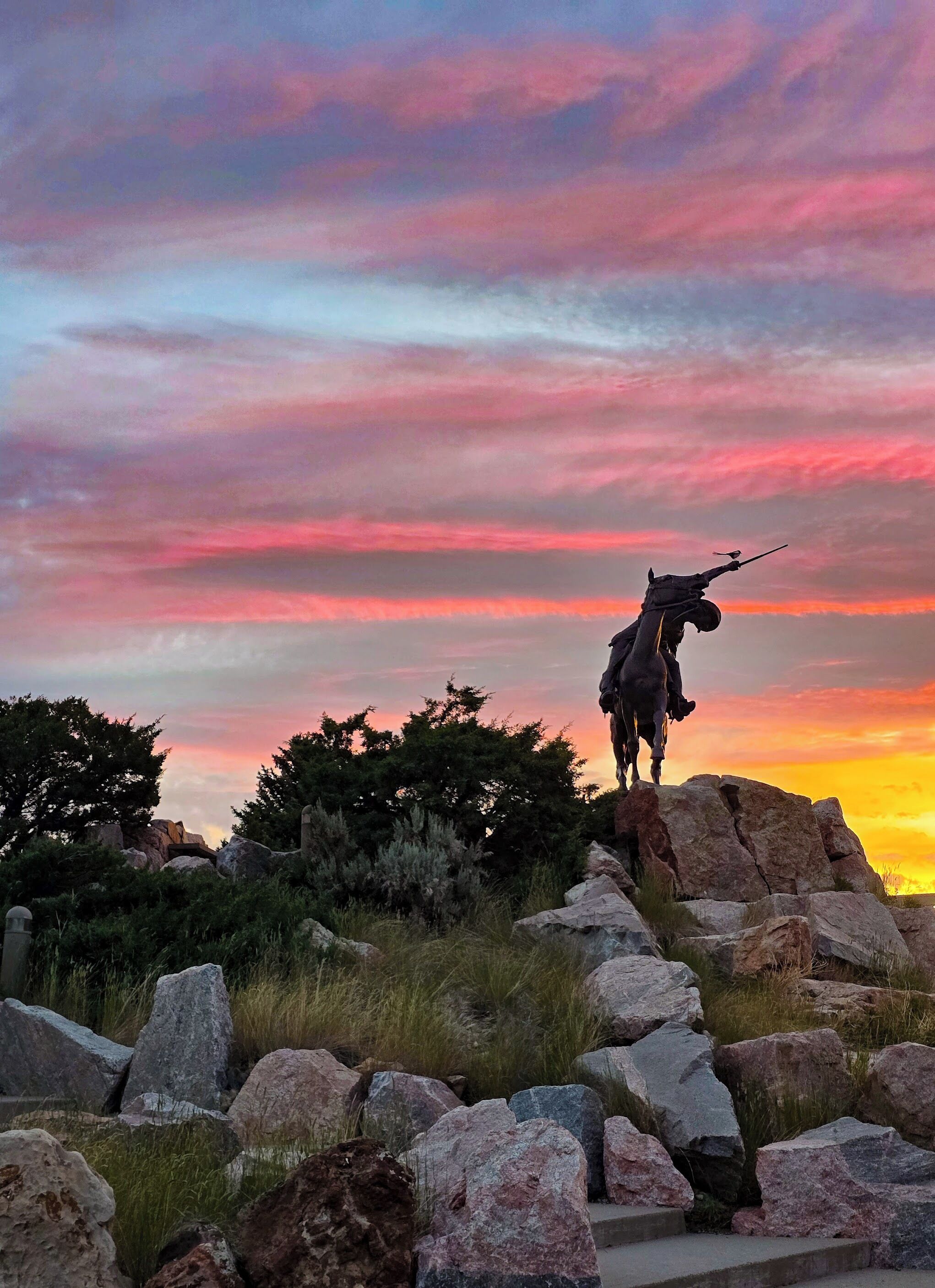 A statue of a person with a rifle on a rock formation with a sunset in the background.