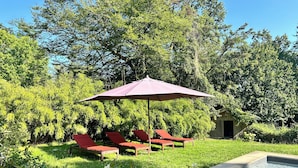 Un espace piscine agrémenté de chaises longues rouges et d’un grand parasol, le tout entouré d’une végétation luxuriante et d’arbres.