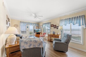 A living room with a ceiling fan, a television, and a view of the ocean through the windows.
