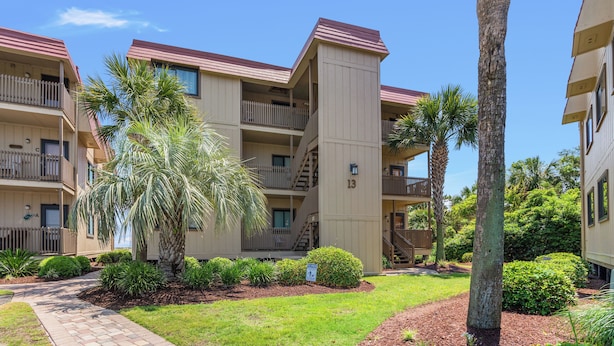 A multi-story residential building with balconies, a paved walkway, and landscaped gardens including palm trees and shrubs.