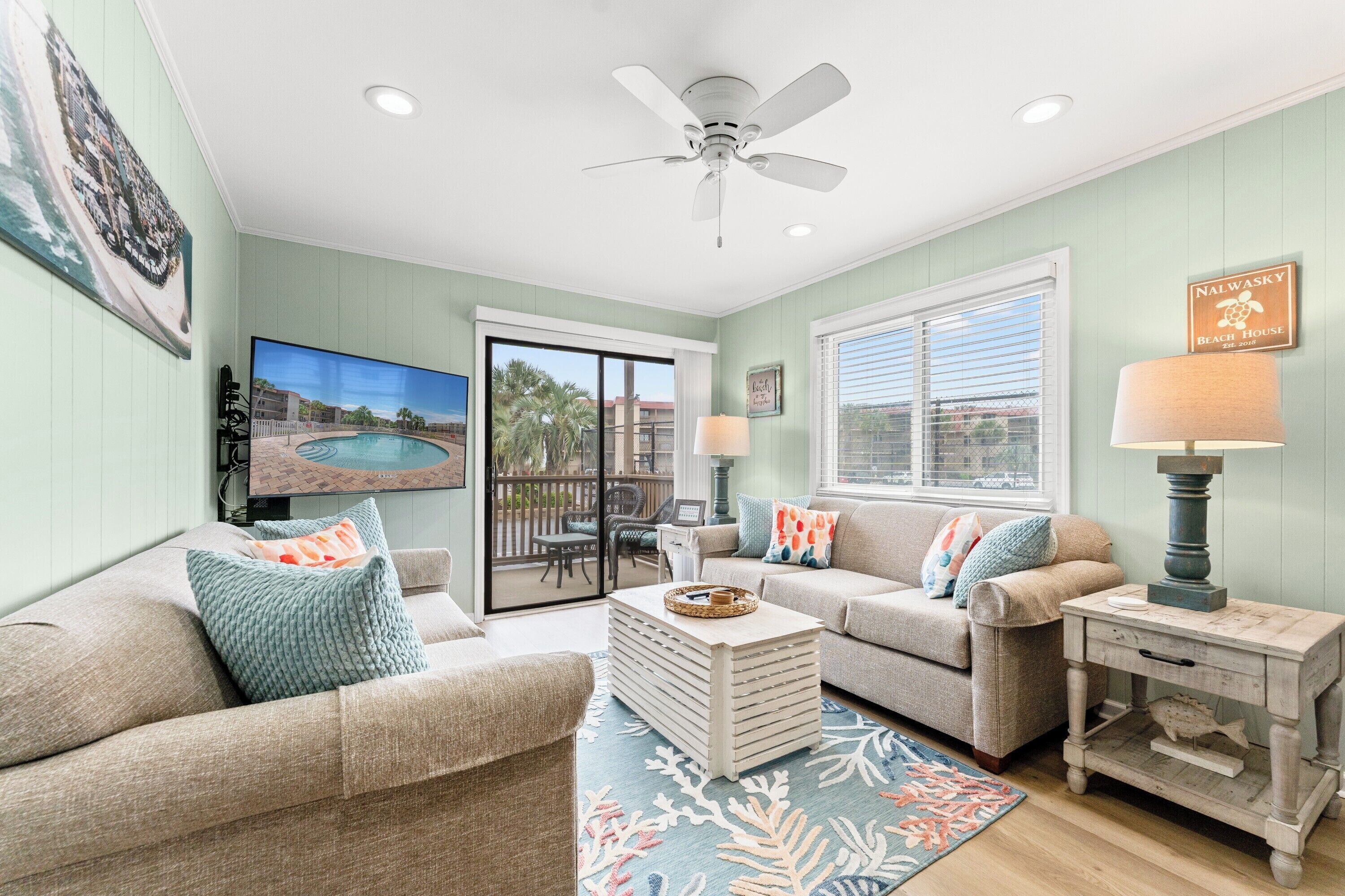 A living room with a pool view, a ceiling fan, and a beach-themed sign.
