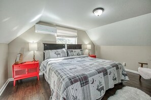 A bedroom with a bed, a red nightstand, a white chair, and an air conditioning unit.