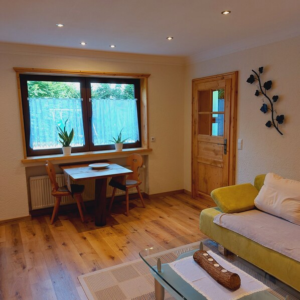 A cozy room with a wooden floor, a dining table with chairs, a sofa, a radiator, and a window with lace curtains.