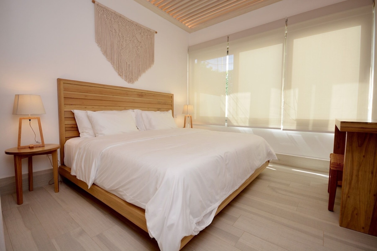 A neatly arranged bedroom with a wooden bed, two bedside tables with lamps, a macrame wall hanging, and a large window with sheer curtains.