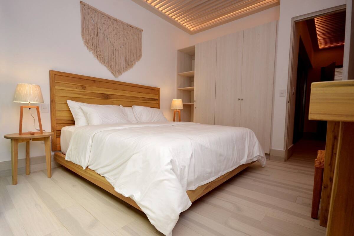 A bedroom with a wooden bed, two bedside tables with lamps, a wooden wardrobe, and a macramé wall hanging.