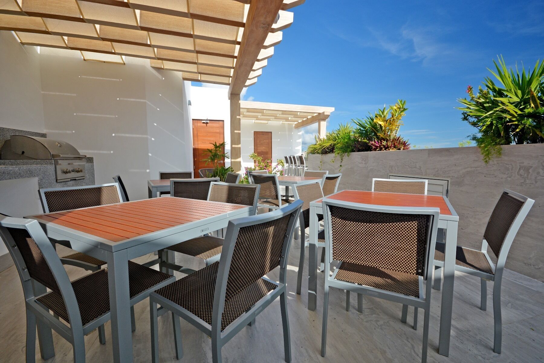 An outdoor patio with tables and chairs, a grill, and a view of the sky and plants.