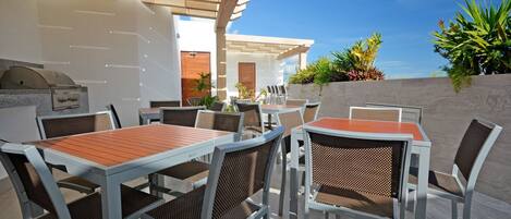 An outdoor patio with tables and chairs, a grill, and a view of the sky and plants.