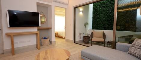 Una sala de estar moderna con un televisor de pantalla plana, una mesa de centro de madera, un sofá gris y un ventanal con una pared verde en el exterior.
