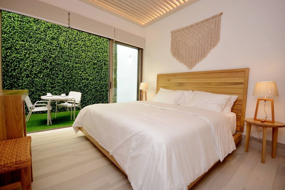 A bedroom with a large bed, a wooden headboard, two bedside tables with lamps, a view of a green hedge through a sliding door, and a small table with chairs outside.