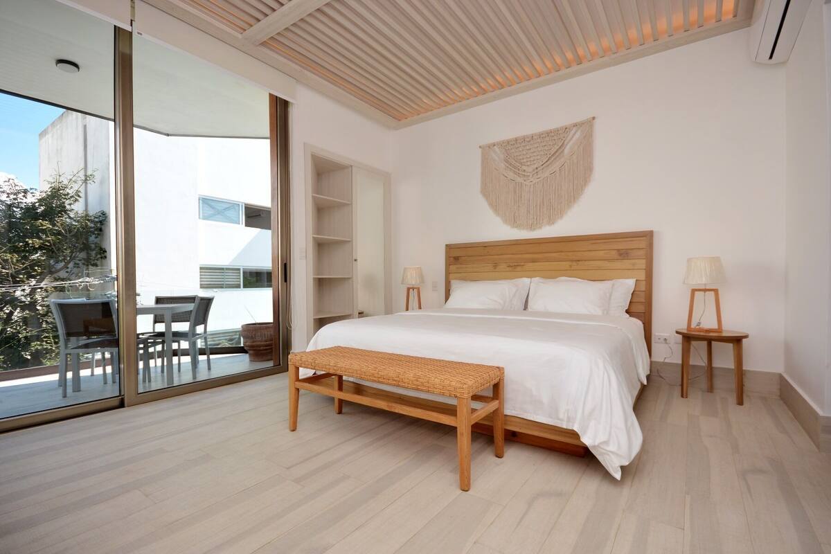 A modern bedroom with a wooden bed, a bench, and a glass door leading to a balcony with outdoor furniture.