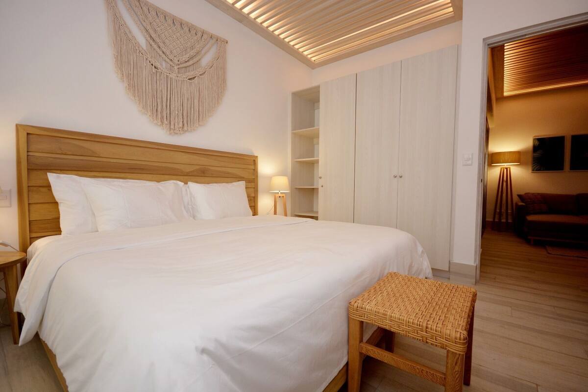 A bedroom with a wooden headboard, a bed with white linens, a woven bench, a macrame wall hanging, and a wardrobe with open doors.