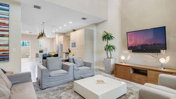 A modern living room with a white coffee table, grey sofas, a flat-screen TV, a potted plant, and a kitchen in the background.
