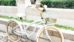 Uma bicicleta branca com detalhes dourados estacionada em um caminho de paralelepípedos ao lado de um prédio com fachada branca e varanda.
