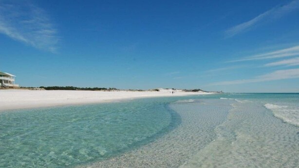 Uma praia com águas claras e rasas, areal arenoso e um edifício à esquerda.