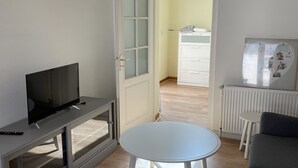 Ein modernes Wohnzimmer mit Fernseher auf einer grauen Unterhaltungsecke, einem weißen runden Tisch, einem grauen Sofa und einem großen Fenster mit Blick ins Freie.