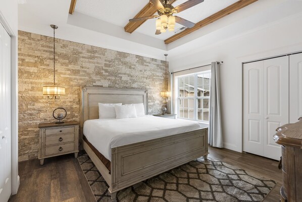 A bedroom with a stone accent wall, a wooden ceiling fan, a bed with white linens, and a nightstand with a lamp.