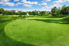 Un campo de golf con un mástil de bandera, césped cuidado y edificios residenciales al fondo.