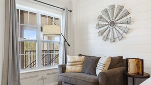 A living room with a grey sofa, patterned curtains, a wall-mounted clock, and a window with a view of a balcony.