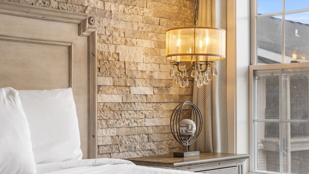 A neatly made bed with white linens, a stone-textured wall, a modern chandelier, and a window with a view.