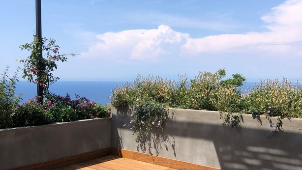 An outdoor patio with wooden flooring, two grey cushioned seating areas, and a view of the sea. - new Apartment - Ischia Garden Suites (Forio)