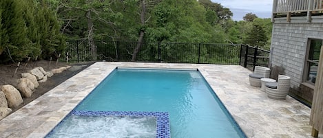 A rectangular swimming pool with a hot tub in the center, surrounded by stone tiles and a black metal fence.
