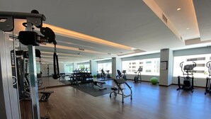 A spacious gym with various exercise equipment, large windows, and a wooden floor.