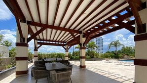 An outdoor patio with a covered seating area, a pool, and a view of palm trees and a building in the background.