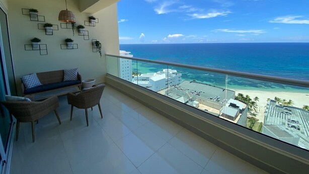 A balcony with a view of the ocean and beach, a sofa, two chairs, and wall-mounted planters.