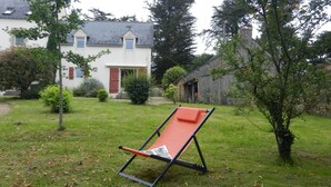 Un jardín con una silla reclinable roja, una casa blanca con puerta roja y un edificio de piedra al fondo.