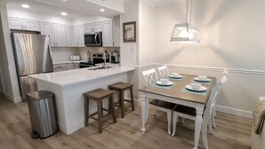 A modern kitchen with white cabinetry, a stainless steel refrigerator, a microwave, and a coffee maker. There is a dining area with a wooden table and white chairs.