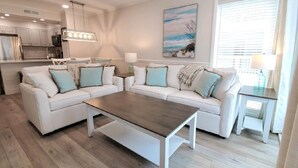 A modern living room with a white sofa set, a wooden coffee table, and a kitchen in the background.