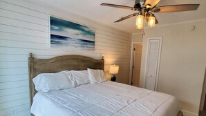 A bedroom with a wooden headboard, a white bedspread, a ceiling fan, and a painting of a beach scene.