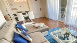 A modern living room with a beige sectional sofa, blue and white cushions, a glass coffee table with a vase, and a dining area in the background.