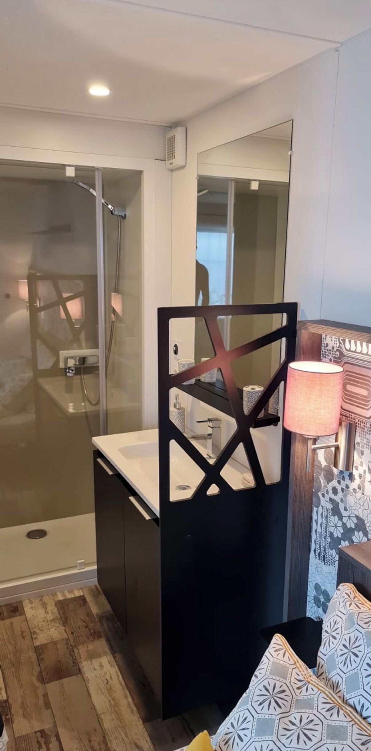 Une salle de bain moderne avec une douche à parois de verre, un meuble-lavabo noir avec un miroir et des coussins à motifs sur un plancher en bois.