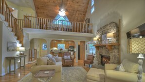 A spacious living area with a high ceiling, wooden staircase, and a stone fireplace.
