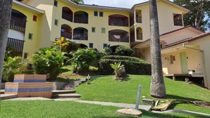 A multi-story yellow building with balconies, surrounded by lush greenery and palm trees.