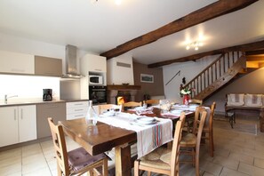A dining area with a wooden table set for four, surrounded by chairs. A fireplace is visible in the background, and a staircase leads to an upper level.