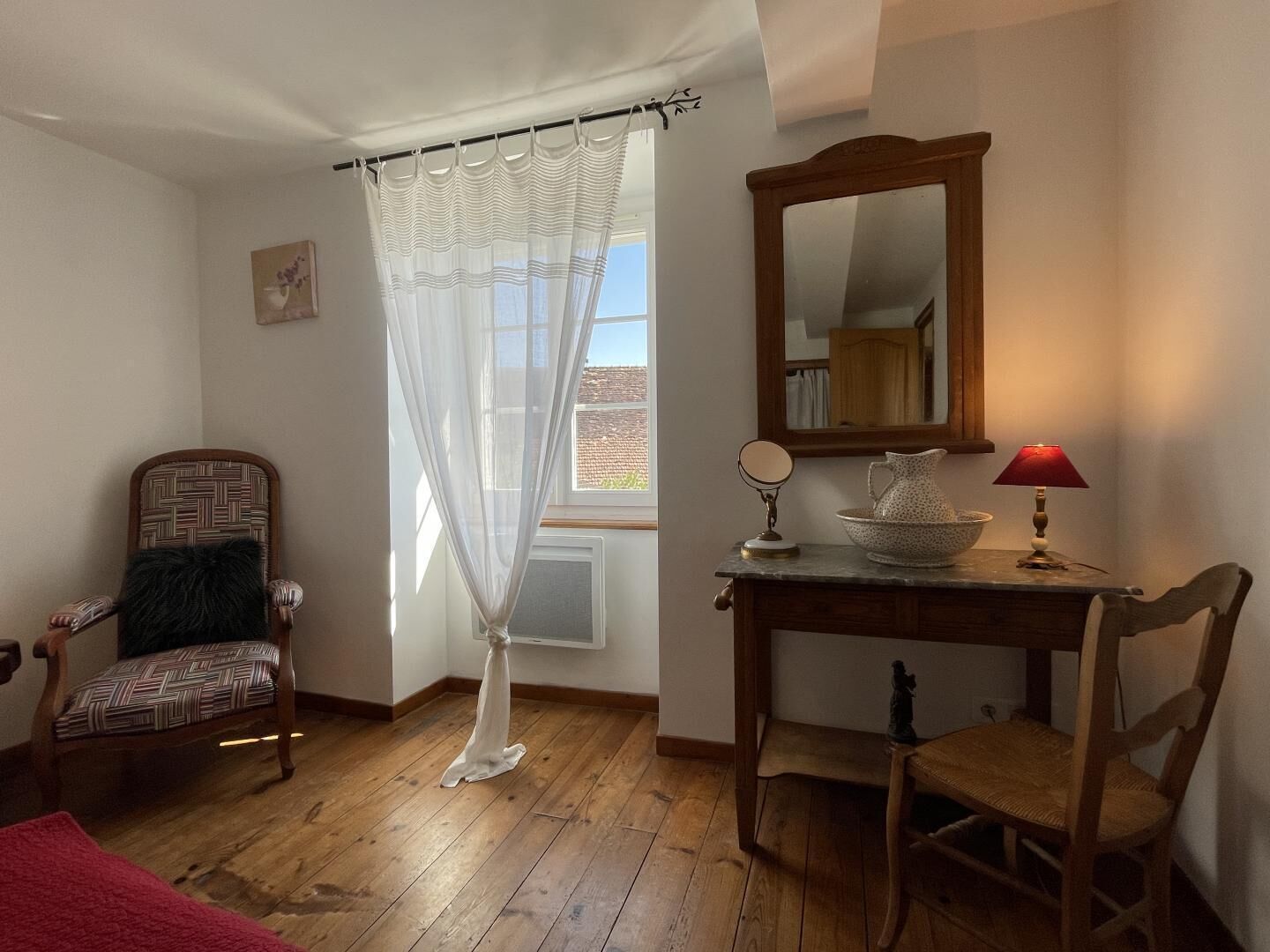 A room with a wooden floor, a chair, a table with a lamp, a mirror, and a window with a curtain.