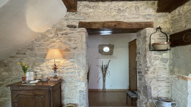 Une pièce rustique aux murs de pierre, avec un plafond en bois, une lampe posée sur une table en bois, un vase rempli de fleurs et un banc en pierre.